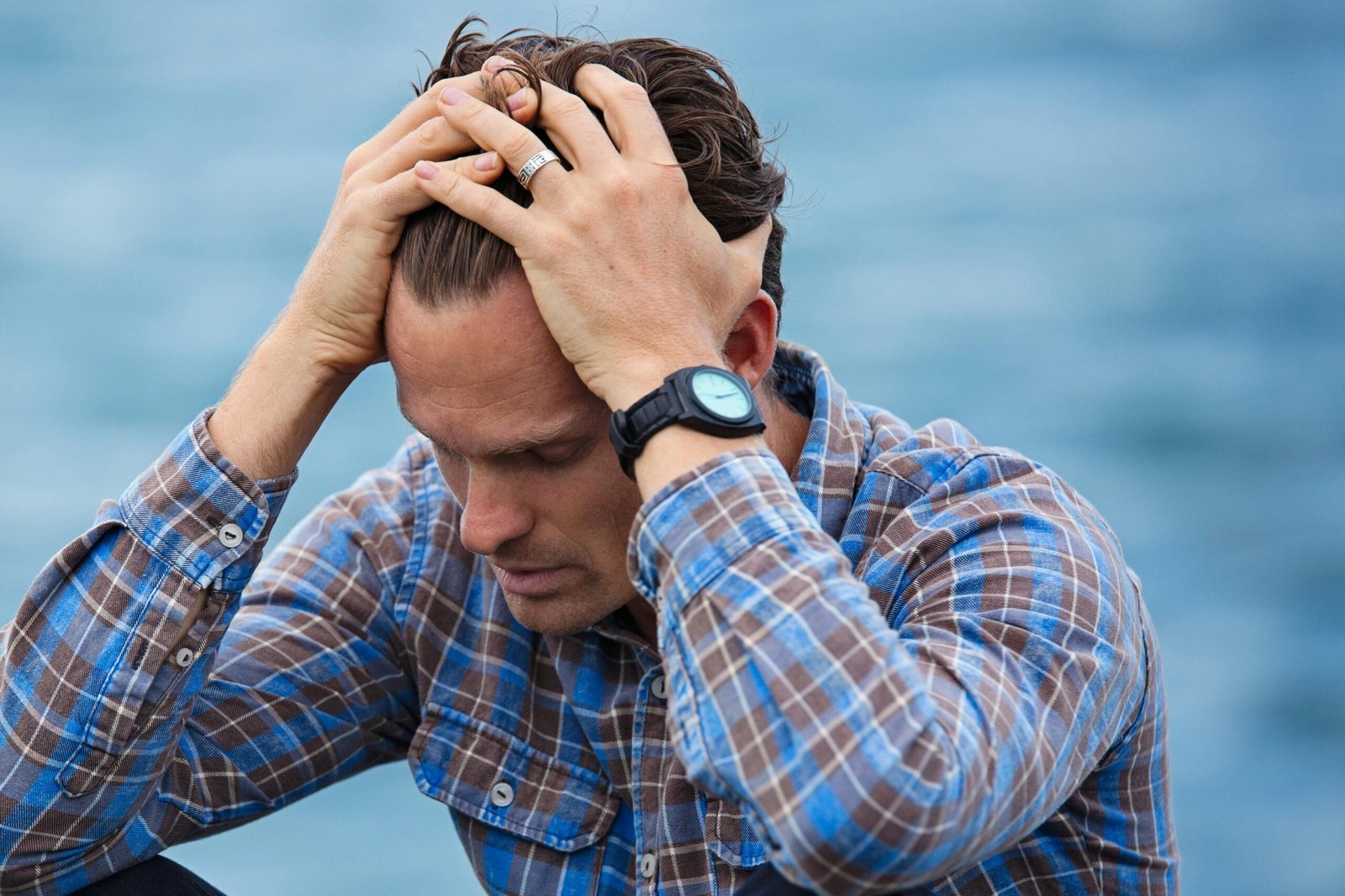 A man in a plaid shirt sits by the water looking distressed, symbolizing stress.